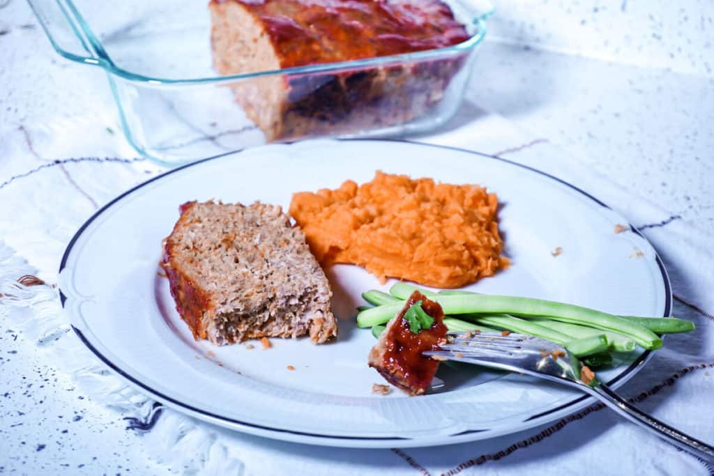 A white plate with a slice of meatloaf, a small serving of mashed orange sweet potatoes and some green beans on a side. On the right side the fork with a bite of meatloaf resting on the plate.