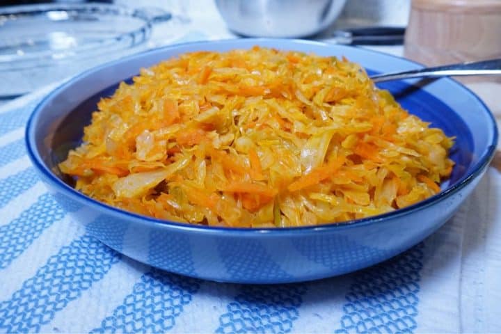 Cooked shreds of cabbage and carrots in a blue plate. The plate is placed on a striped blue and white kitchen towel. 