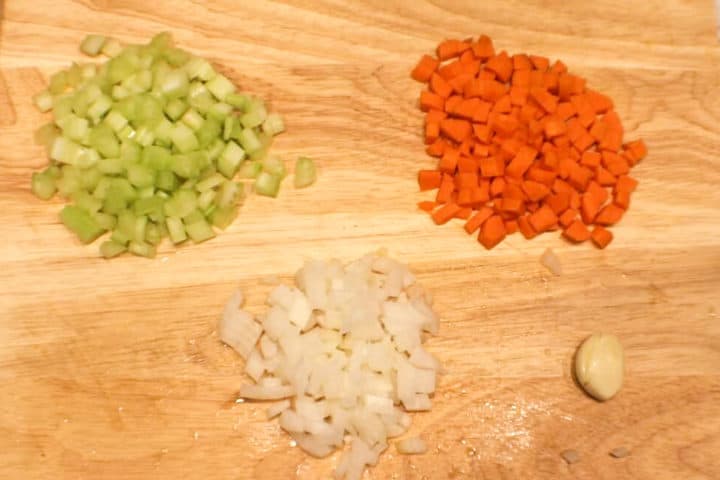On a cutting board there are finely diced carrots, celery and onions and one peeled garlic clove.