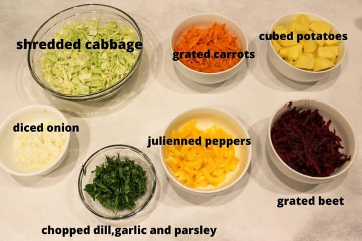 Overhead shot of chopped vegetables to make the recipe: shredded cabbage, diced onion, chopped garlic, dill and parsley, grated carrots and beets, julienned sweet peppers and cubed potatoes. All these vegetables are in separate bowls and are labeled. 