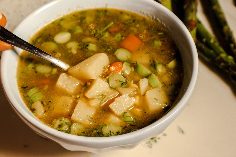 A blue bowl with a chunky yellow and a hand holding a spoon submerged in the soup on the left side. There is visible chunks of potatoes, sliced asparagus, carrots and green dill in the soup.