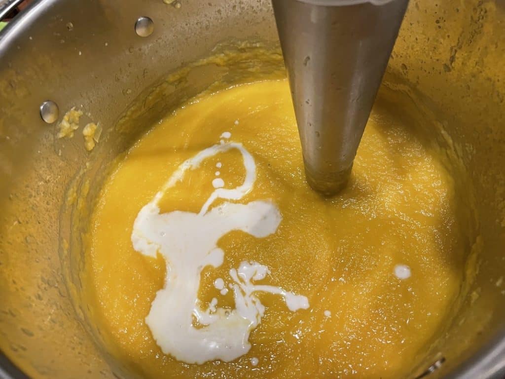 A pot with already blended creamy yellow soup and just added splash white milk on the surface. There is a immersion blender submerged in a soup. 