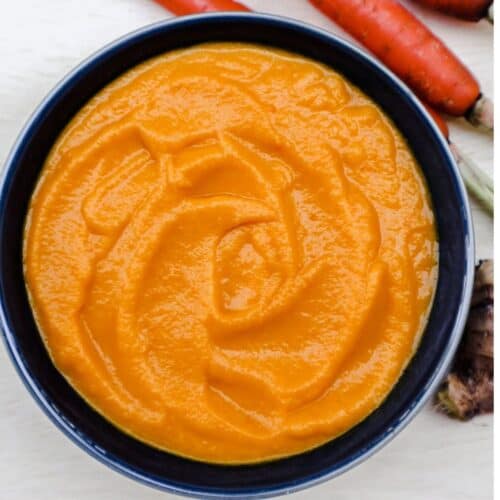 A dark blue bowl with creamy orange carrot soup and visible fresh carrots in the top right corner.