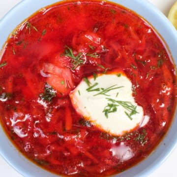 Overhead shot of blue bowl filled with red borscht soup topped with a dollop of sour cream and some chopped fresh dill on top.