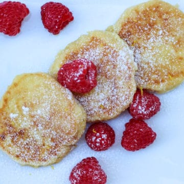 hree syrniki pancakes served on a white plate, topped with powdered sugar and three syrniki pancakes served on a white plate, topped with powdered sugar and raspberries