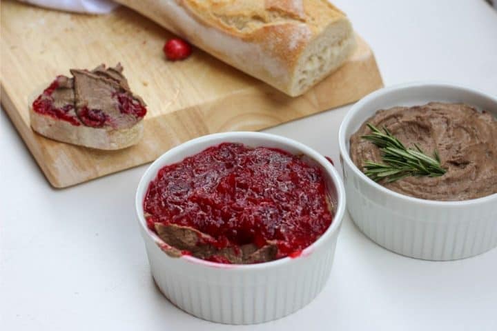 close up shot of chicken liver pate in a jar, topped with cranberry sauce. You can see a frehs cut baguette in the back on the cutting board and a slice topped with the liver spead. 