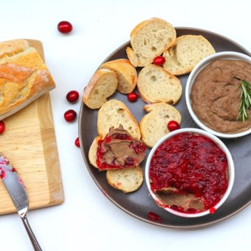 Fresh cut bagette on a cutting board. Next to the baguette, there is a dark grey shallow plate with slices of the bagette, one of which topped with chicken liver pâté. On the same plate, there is 2 small jars with chicken liver spread: one has cranberry sauce on top, another one has rosemary.