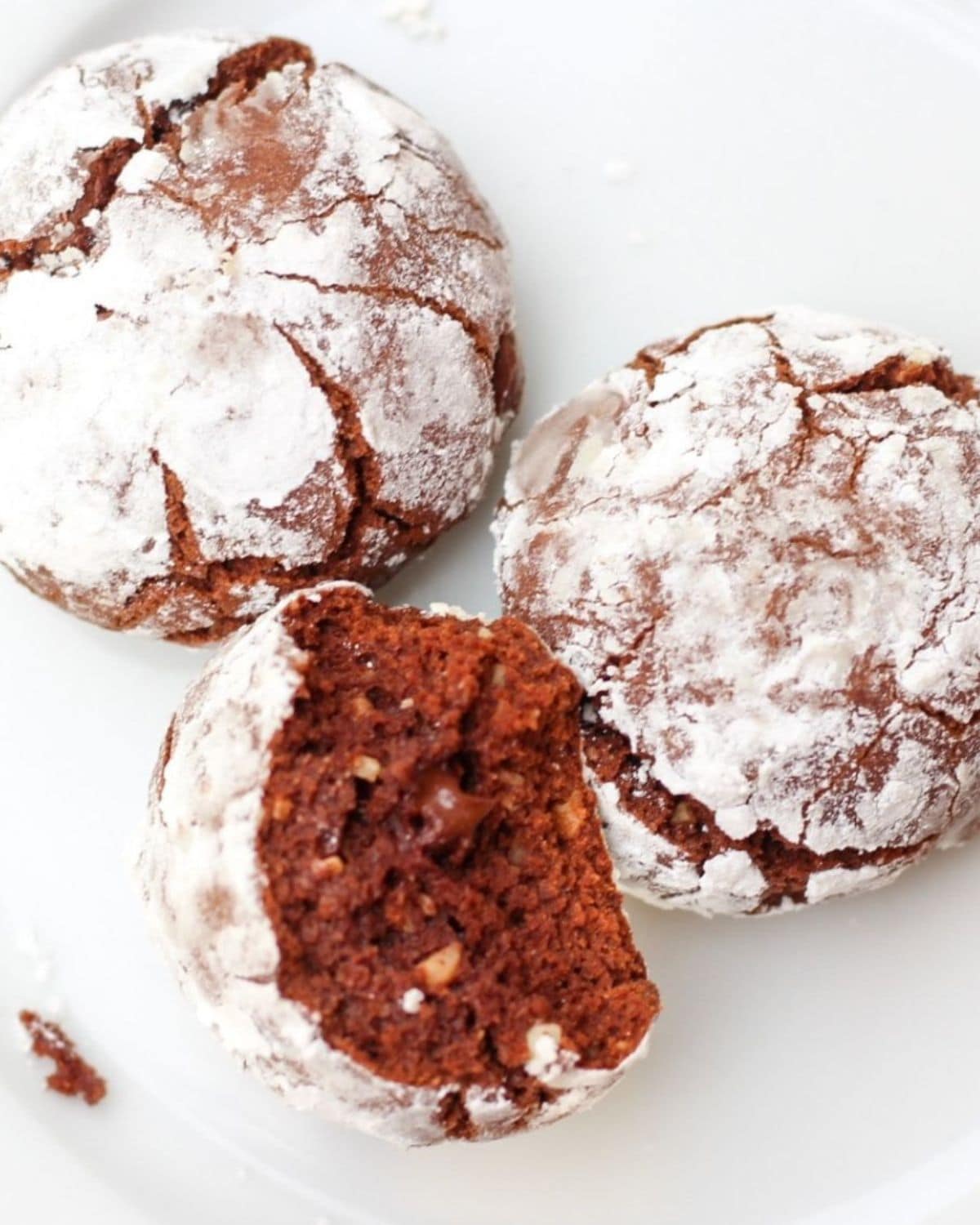 Three chocolate crinkle cookies on a white plate. One of cookies is halved showing the chocolate soft texture in the inside.