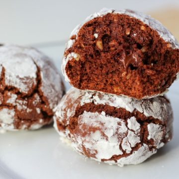 A plate with three chocolate crinkle cookies, two of them are on top of each other. The top cookie is broken in half to show the inside with chocolate dough and melted chocolate chips.