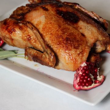 Whole cooked duck on a large white serving plate with quarter of pomegranate on corner.