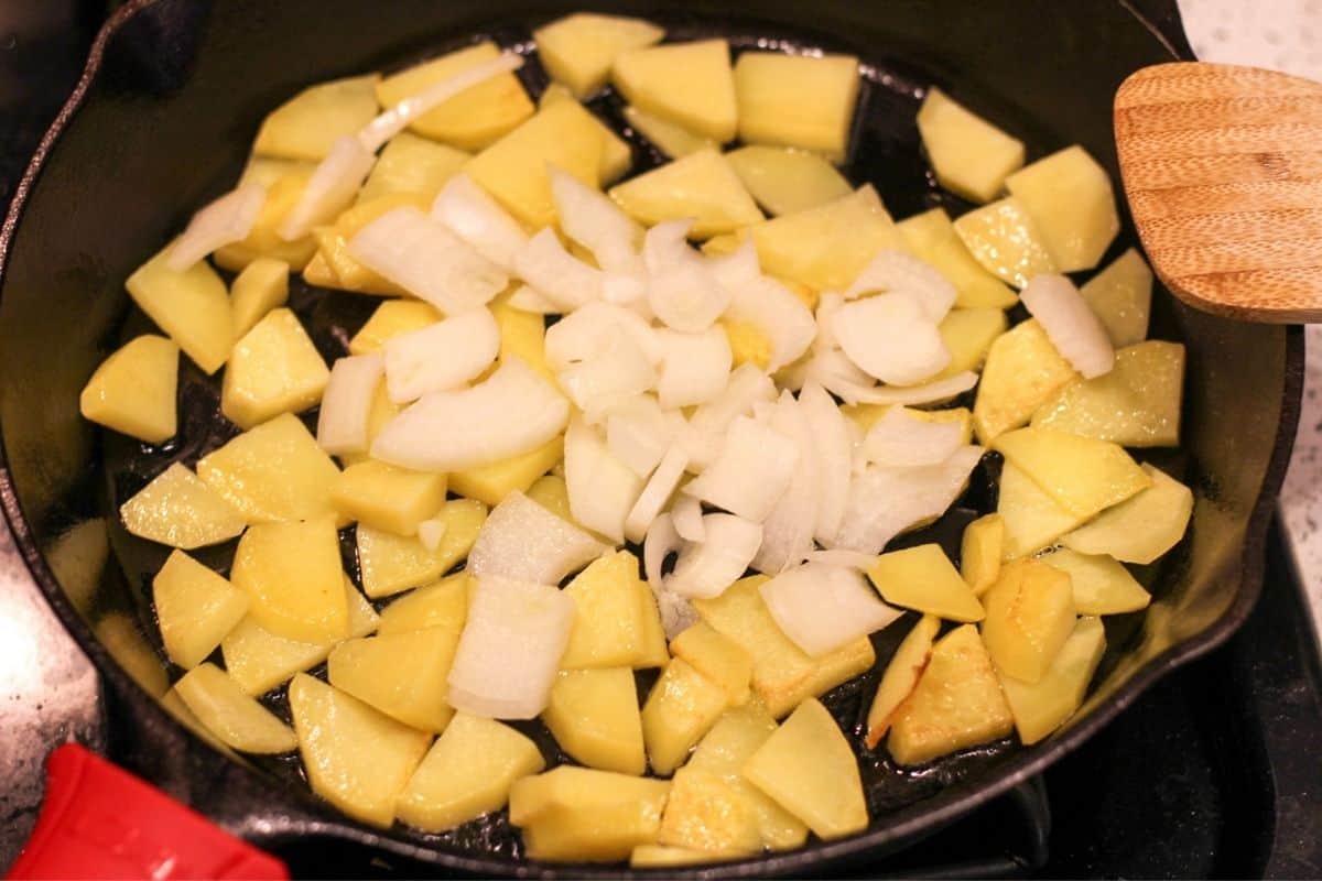 Sliced potatoes are frying in the large pan, sliced onions added on top of potatoes. 
