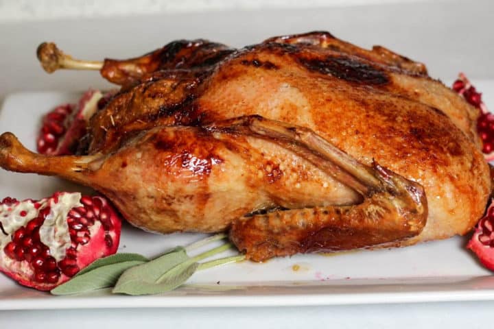 Whole roasted duck on a large white serving plate with quarters of pomegranate on corners
