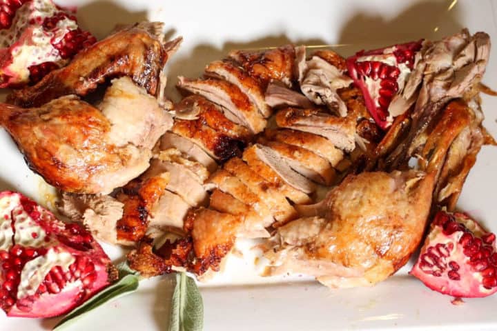 Carved duck meat on a serving plate with some quartered pomegranated on sides and a twine of fresh sage.