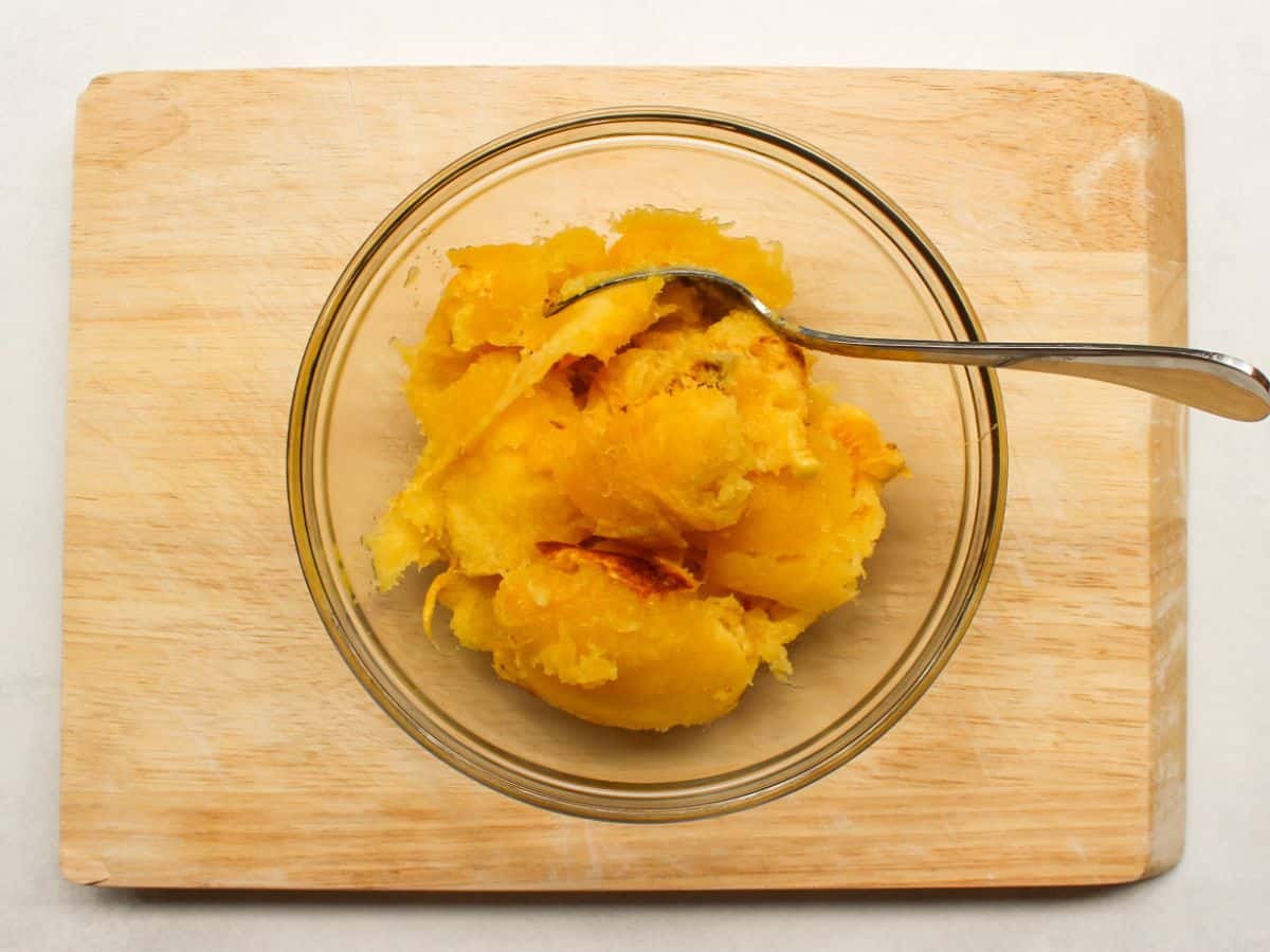 A glass bowl on a cutting board with scooped yellow squash flesh and a spoon.