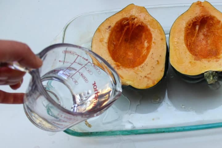 View of cut in half acorn squash in a glass baking dish. Cup of water being poured into the bottom of the dish. 