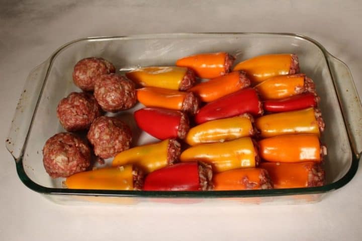 Glass dish with red and orange uncooked stuffed bell peppers and few meatballs on a side from leftover meat.