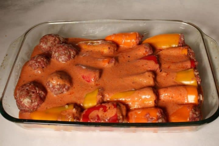 A glass baking dish with stuffed peppers and few meatballs covered in a creamy sauce, ready to go to the oven.