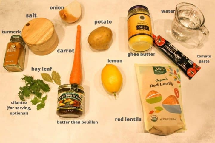 All required ingredients to make the red lentil soup on a flat surface. There are lentils, water, tomato paste, ghee butter, one potato, one carrot, onion, salt, better than bouillon, cilantro, bay leaf and turmeric.