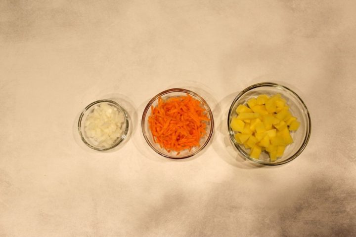 Three bowls with cubed potatoes, shredded carrots and diced onions.