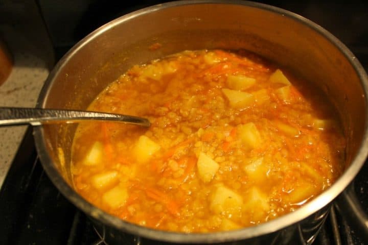 Pot with the cooking lentils, potatoes, carrots, onions and water. There is a spoon submerged in the pot.