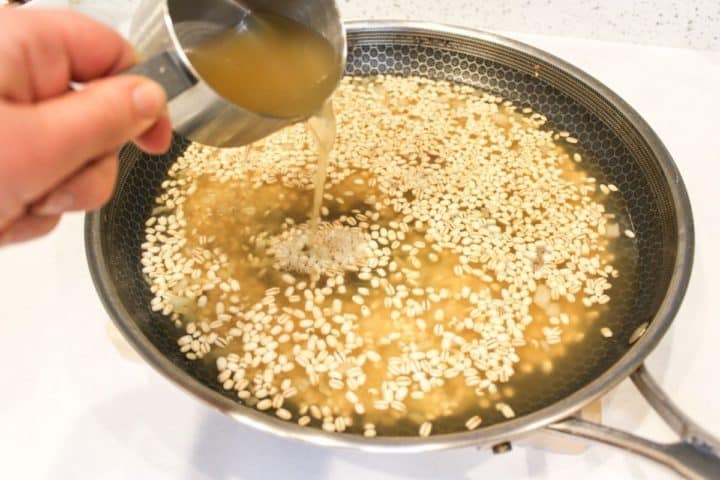 Large frying pan with pearl barley and broth in it, with a cup of more broth being pouring to the pan.