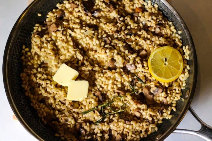 Overhead shot of creamy barley risotto in a frying pan with butter, rosemary leaves and a slice of lemon on top..