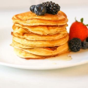 Stacked pancakes on a white plate with some berries on top and on a side.