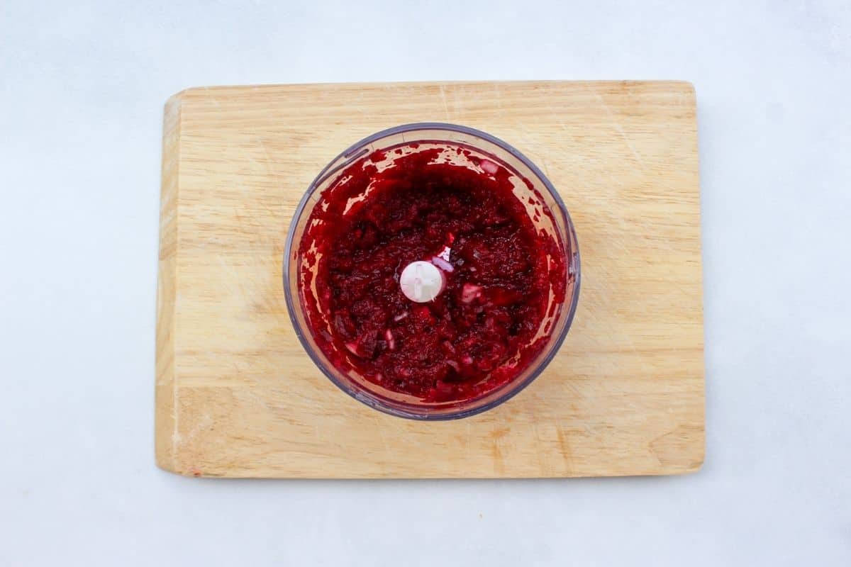 Recipe process shot: a food processor on a wooden cutting board with grated beets and onion.