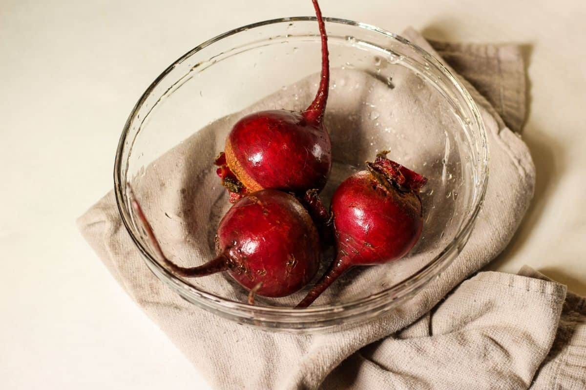 Three washed beets in a clear glass bowl on top of grey kitchen towel.