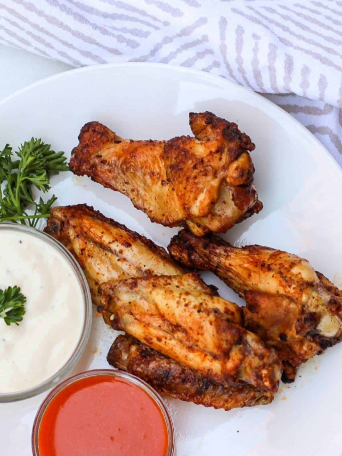 A white plate with 4 cooked wings and two bowl of red sauce and white sauce on a side. there is some fresh parsley on a plate.