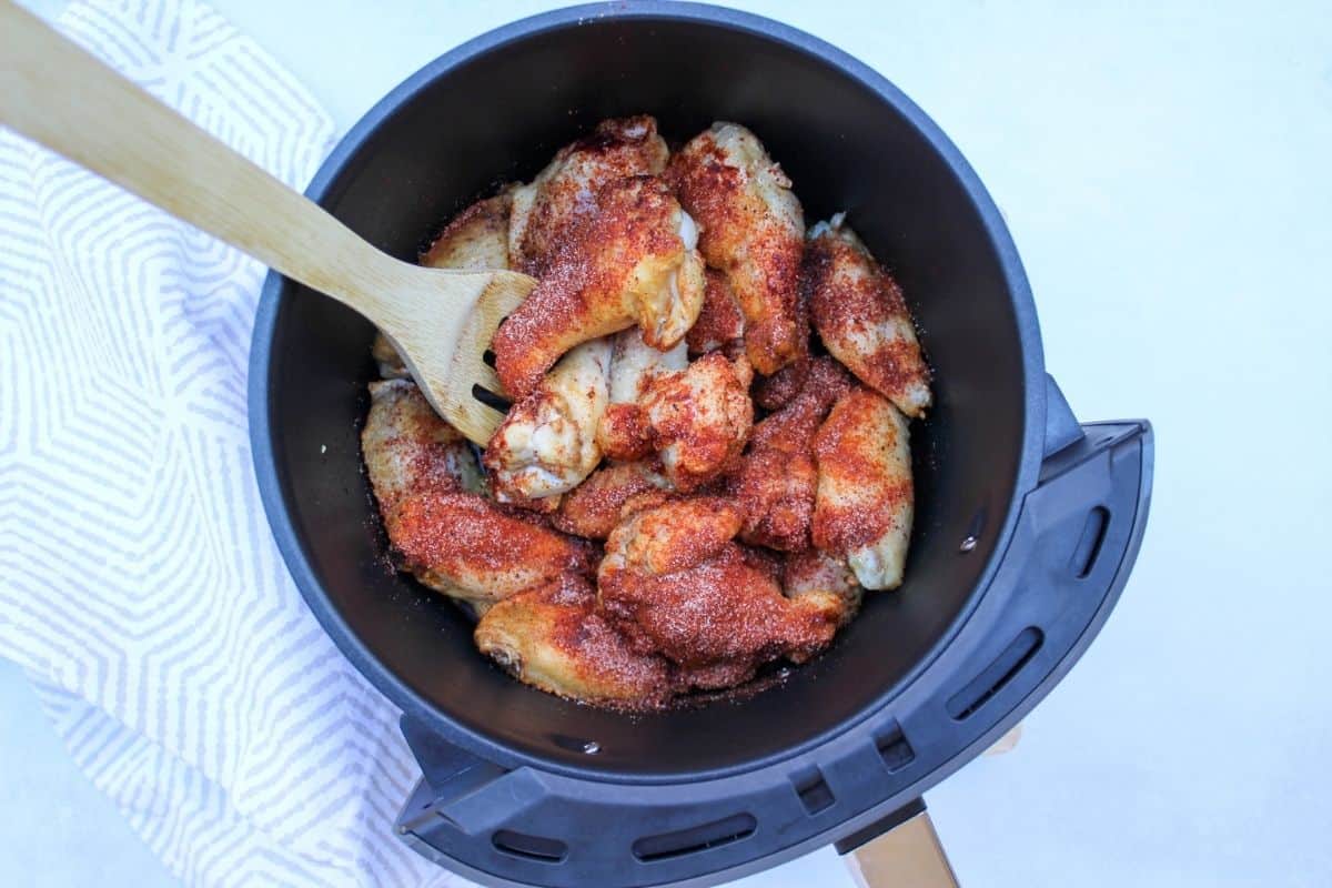 Process shot #2: air fryer basket with hald cooked wings in it and added spices on top. There is a wooden spatula in the air fryer.