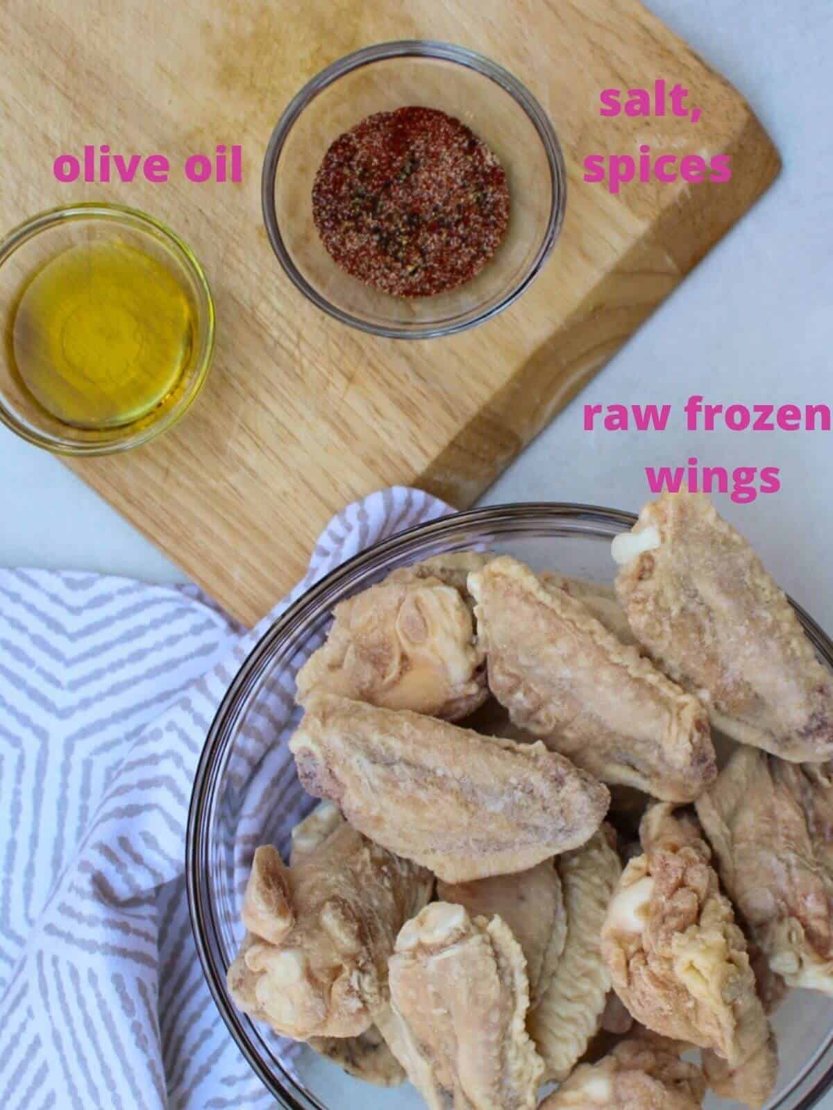 Air fryer frozen wings labeled ingredients: raw frozen wings in a glass bowl , two separate small bowls wil olive oil and spices on a cutting board.