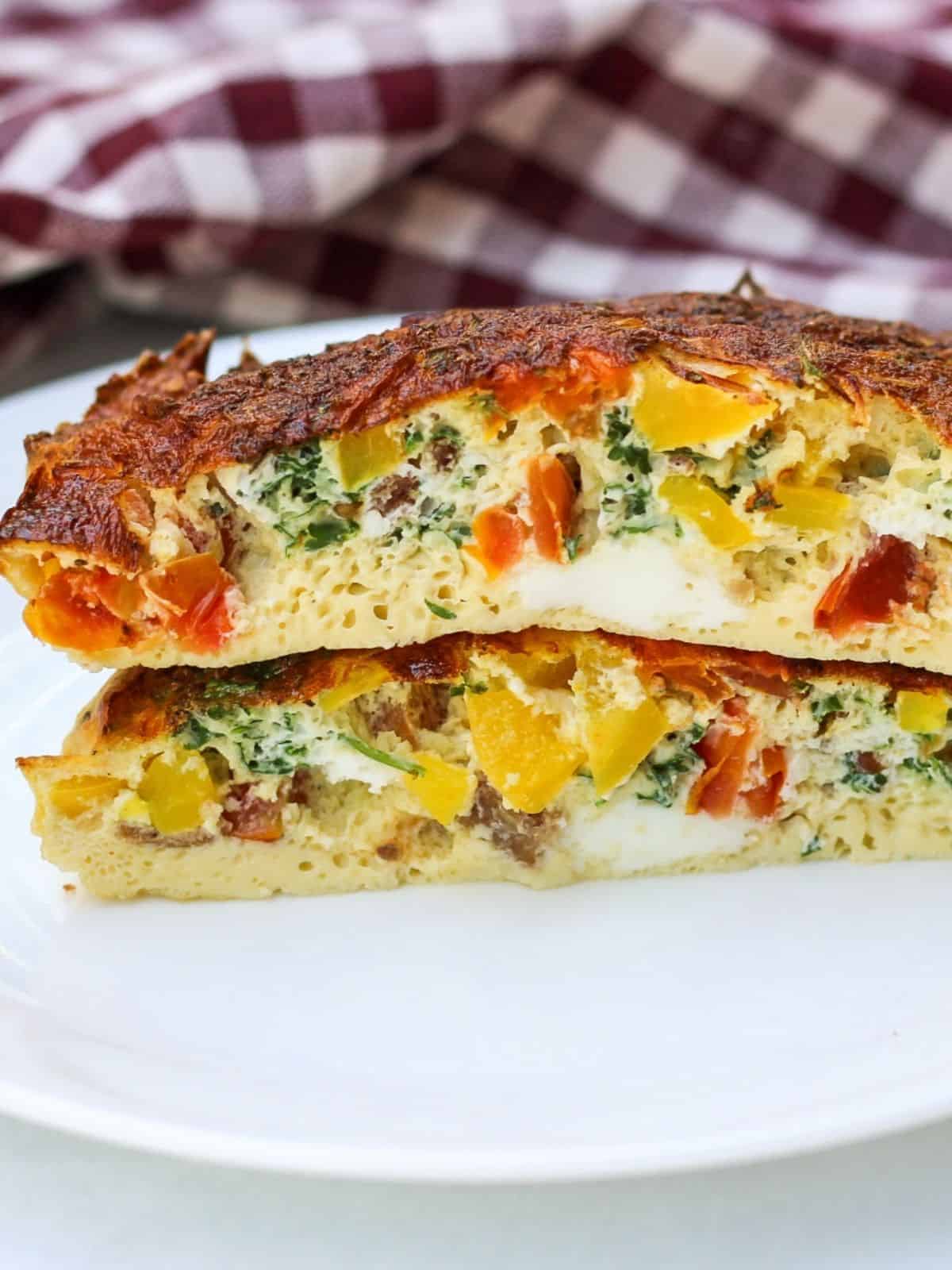 Two large omeltte halves are stacked on each other facing the cut side towards the camera lense. The omelette is fluffy and thick with visible yellow, red and green vegetables inside and golden brown top.