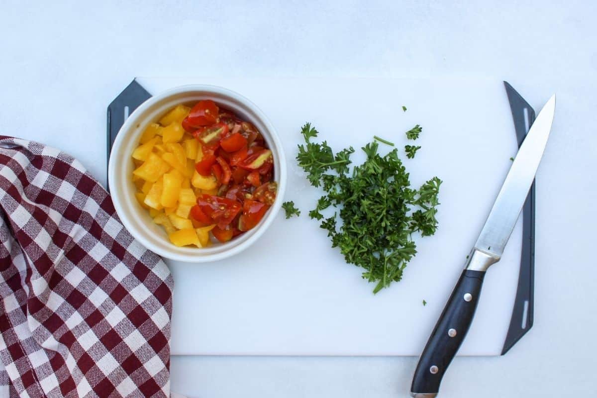 A white board with some chopped fresh parsley. There is also a small bowl on a side with diced tomatoes and yellow bell peppers. The knife is laying on the board on the right side.