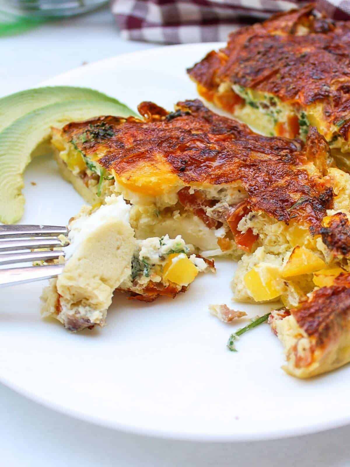 A white plate with omelette. There is a few slices of avocado on a side and a fork with one bite of omelette.