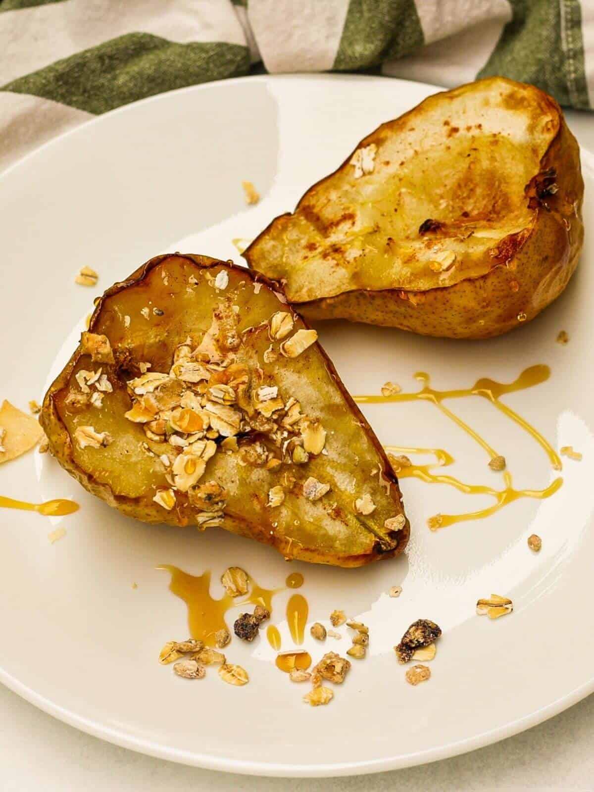 Two baked pear halves facing the cut side up on a white plate. They are drizzles with honey and one of the halves has granola on top.