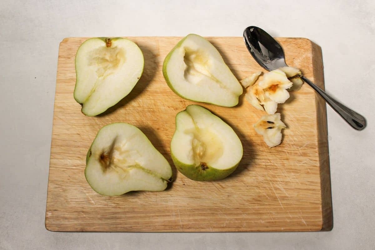 Process shot #2: four pear halves facing the cut side up. The seeds and core has been removed. The seeds and a spoon are on the side of a wooden cuttign board next to the pears.