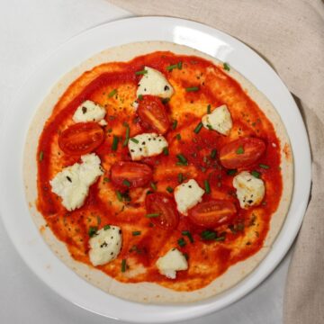 Thin pizza pie on a white plate. Pizza has sauce, crumbles of cheese, tomatoes and spcies on top.