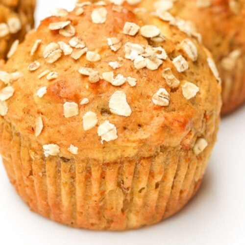 Close-up of moist apple banana carrot muffin with visible oat topping and another muffin blurred in the background.