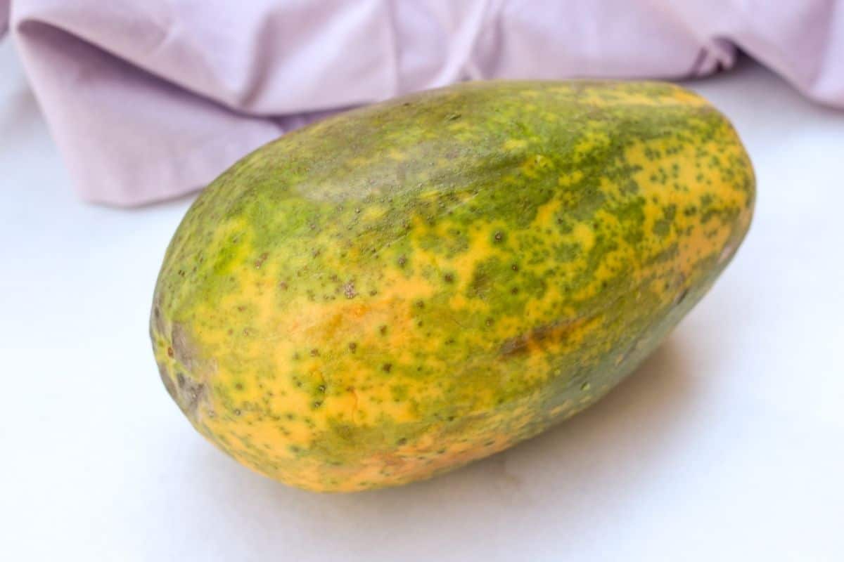 A whole papaya fruit on a white surface with a purple textile behind..