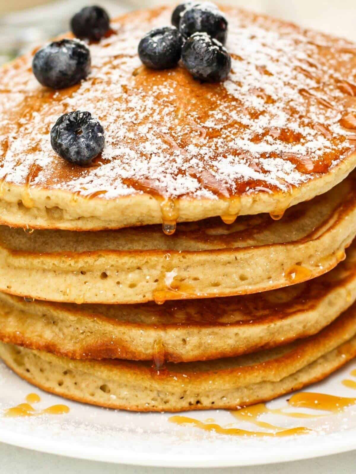 A close up show of a stack of pancakes with some syrup, powdered sugra and blueberies on top.