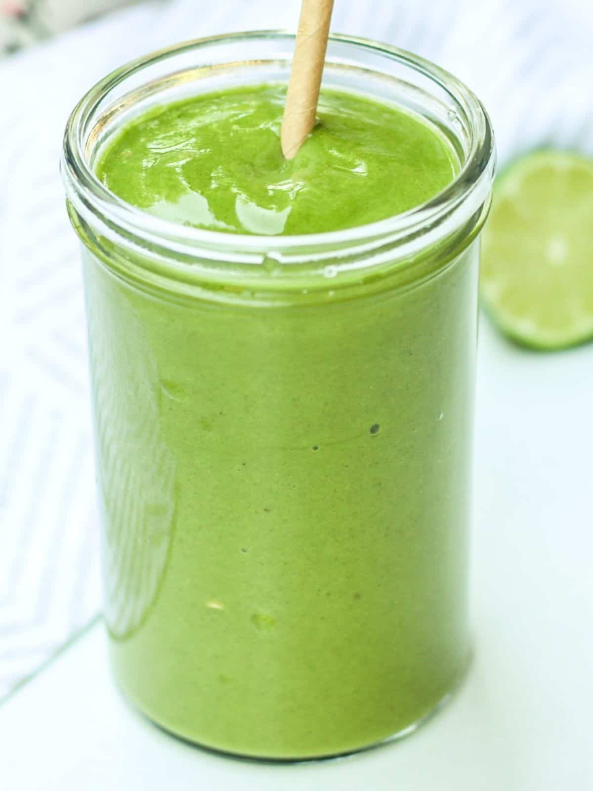 A bright green smoothie in a tall glass with the straw. There is half of the lime behind it. 