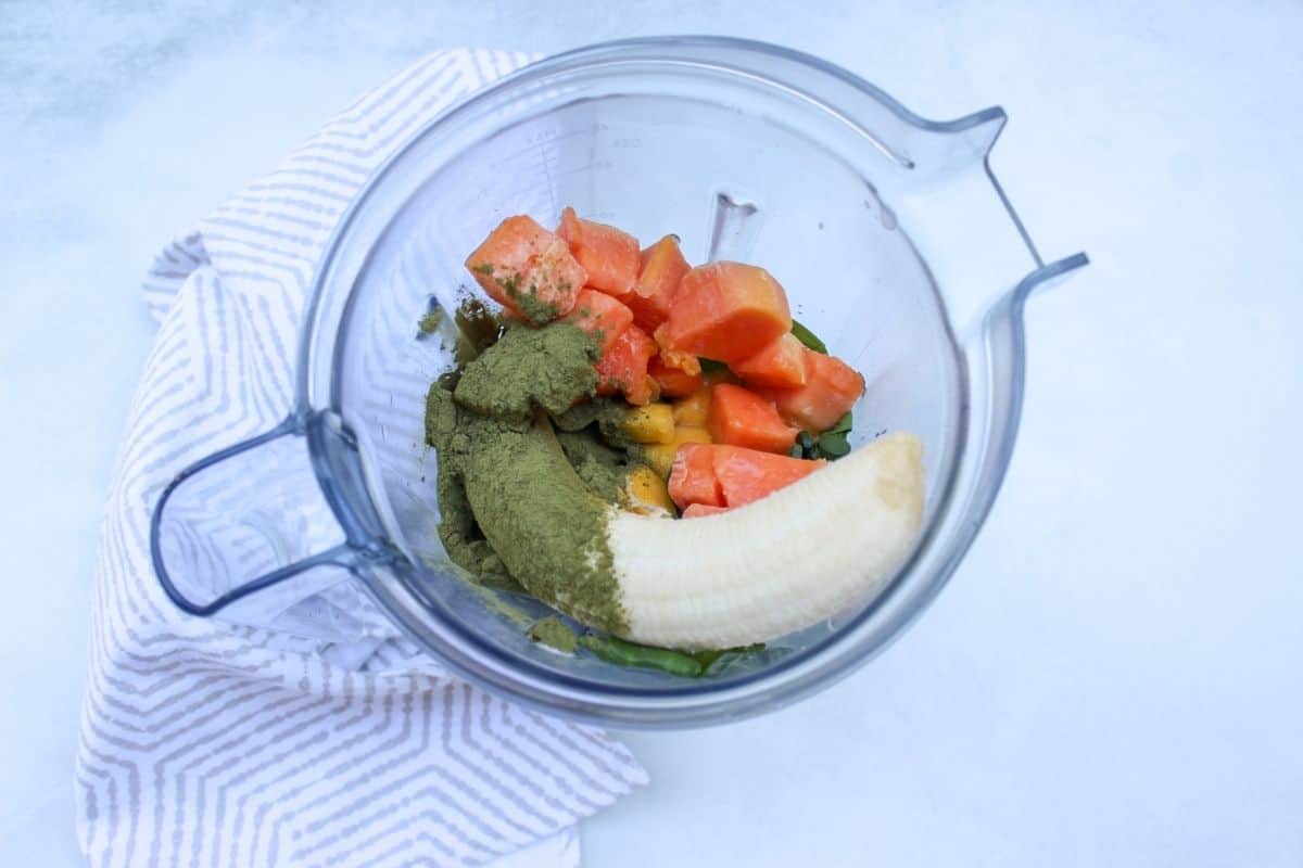 Process shot #1: overhead shot of a vitamix blender container with whole banana, some orange fruits and green powder.