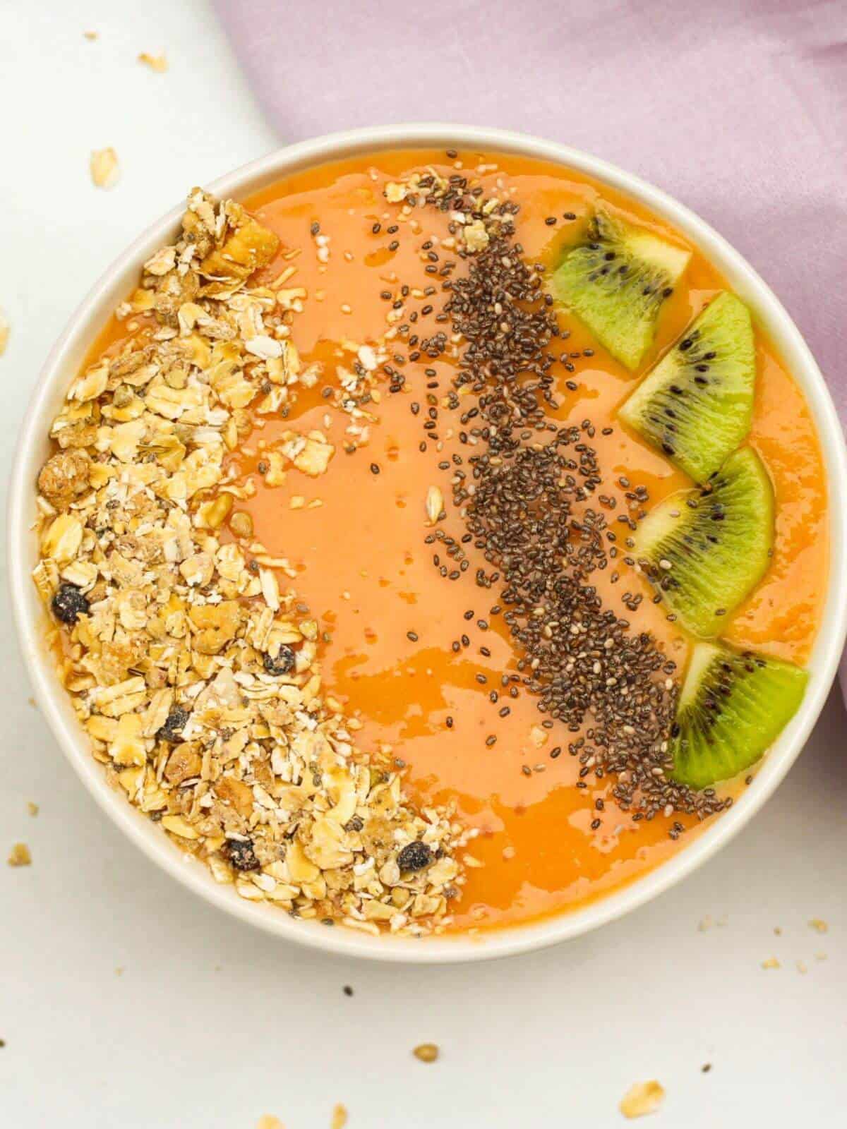 Overhead shot of a white bowl filled with orange papaya smothie. The smoothie is topped with oat muesly, chia seeds, and slices of kiwi fruit. There is a purple towel on the white surface and some sprinkled dry oats all around the white bowl..