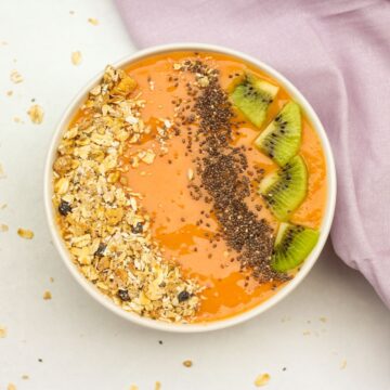 Overhead shot of a white bowl filled with orange papaya smothie. The smoothie is topped with oat muesly, chia seeds, and slices of kiwi fruit. The smoothie is topped with oat muesly, chia seeds, and slices of kiwi fruit. There is a purple towel on the white surface and some sprinkled dry oats all around the bowl.