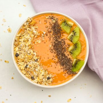 White bowl filled with orange papaya smoothie and topped with oat granola, chia seeds, and slices of kiwi fruit.