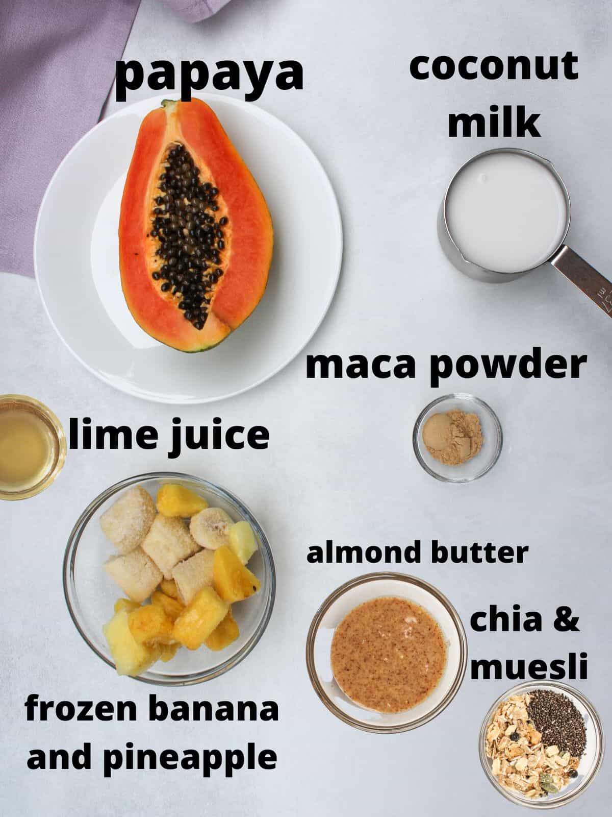 Labeled recipe ingredients on a white background. From top to bottom: half of the fresh papaya with seeds, coconut milk, lime juice, maca powder, frozen bananas, almond butter, chia seeds and muesli.