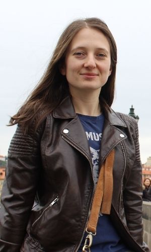 A young woman image from heat to the waist. She has long brown hair and wearing a leather jacket over a dark blue t-shirt. She is smiling and looking at the lens of camera. 