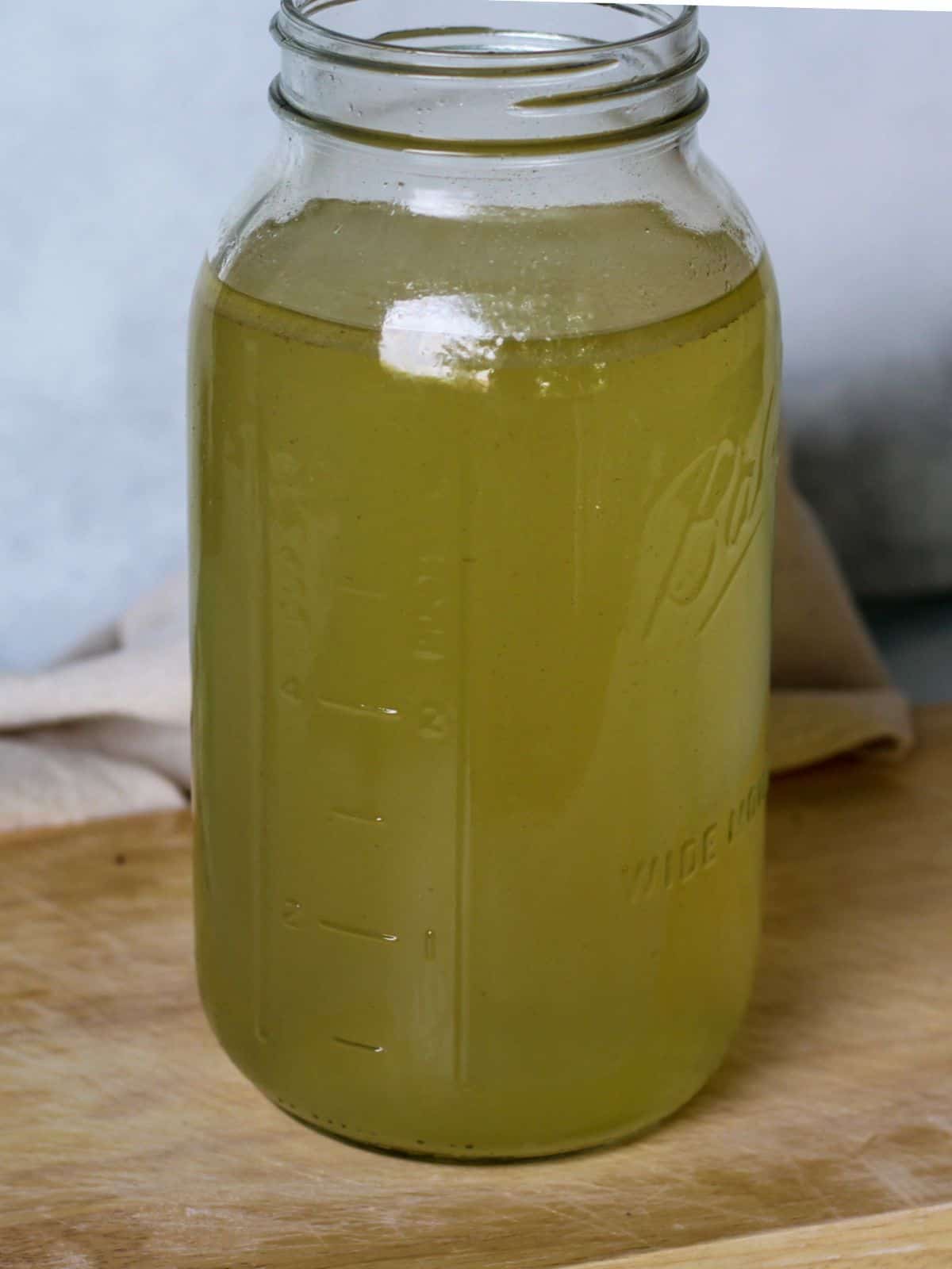 A large glass jar on a wooden cutting board fillied with yellow broth.