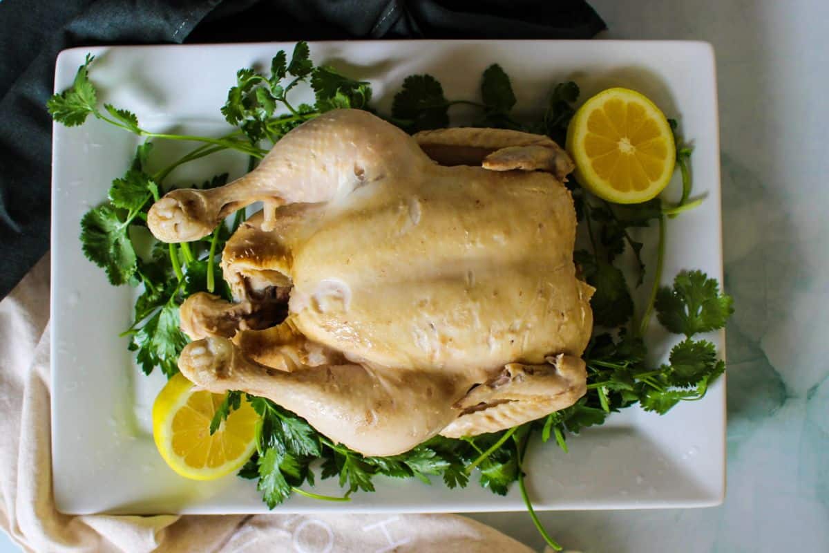 Whole boiled poultry bird placed on a bed of parsley. There are two fresh lemon halves on both sides.